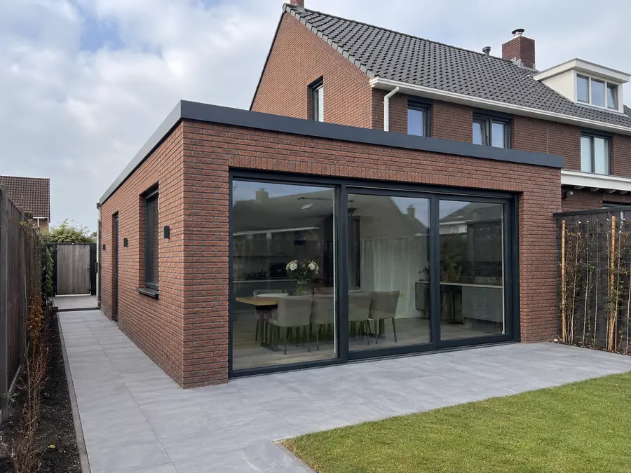Uitbouw met brede schuifpui waardoor de eetkamer zichtbaar is, met terras ervoor.